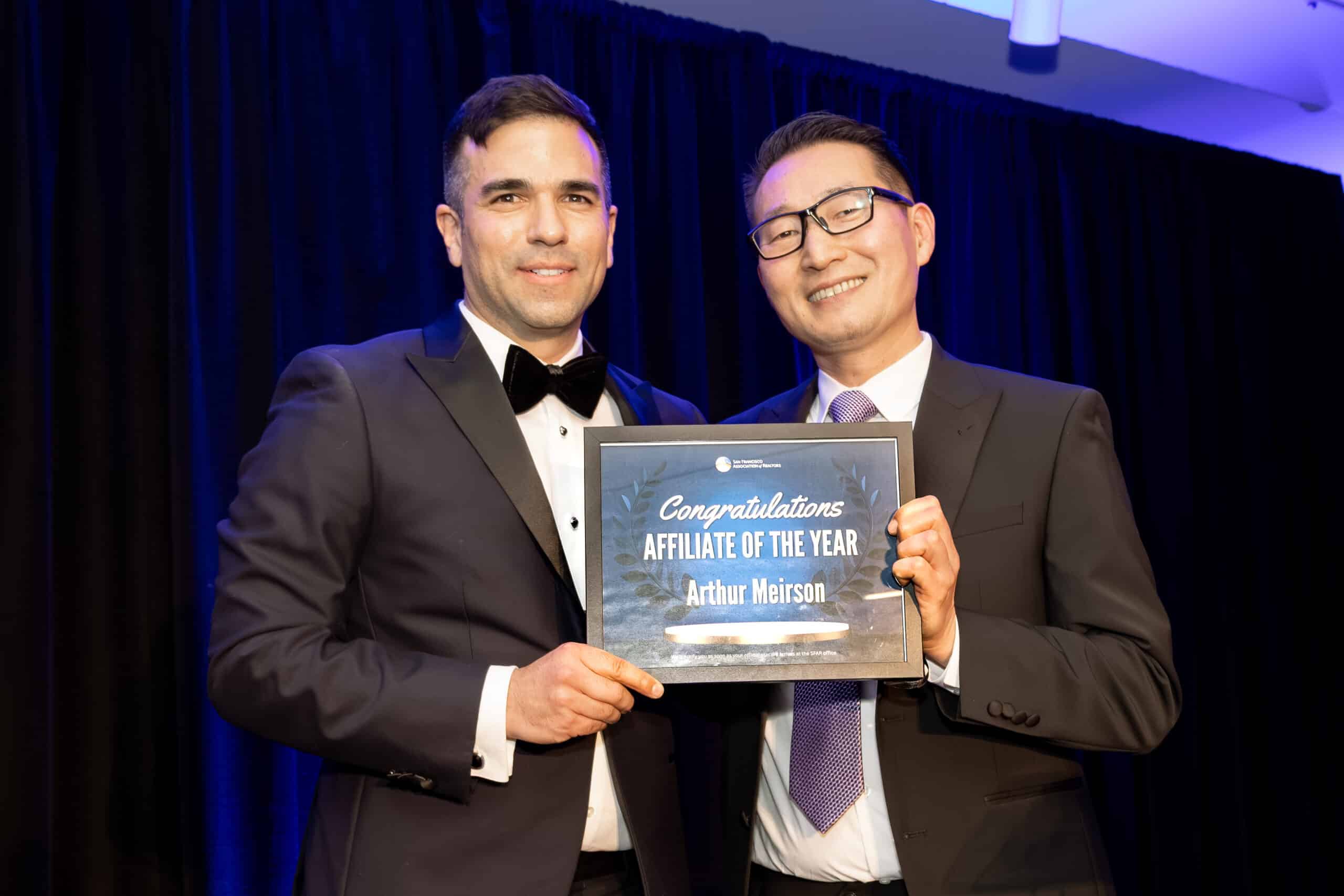 Two men in formal attire holding an award certificate for "Affiliate of the Year" at a professional event, showcasing celebration and recognition in a stylish setting.