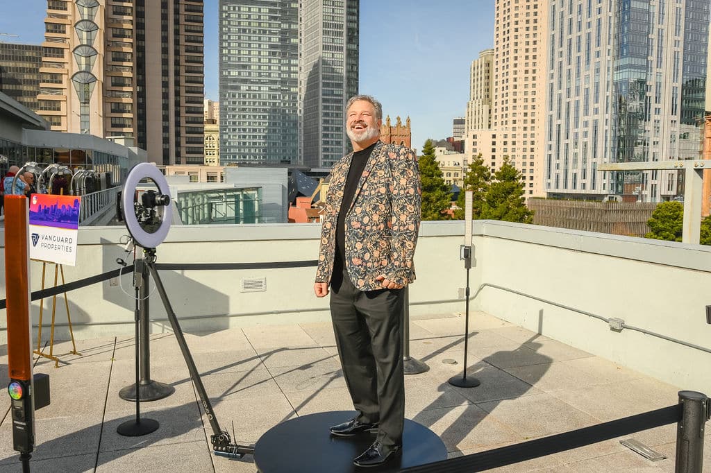 Man posing in a floral jacket at a 360 video booth setup, with a city skyline backdrop, showcasing event marketing and social media engagement.