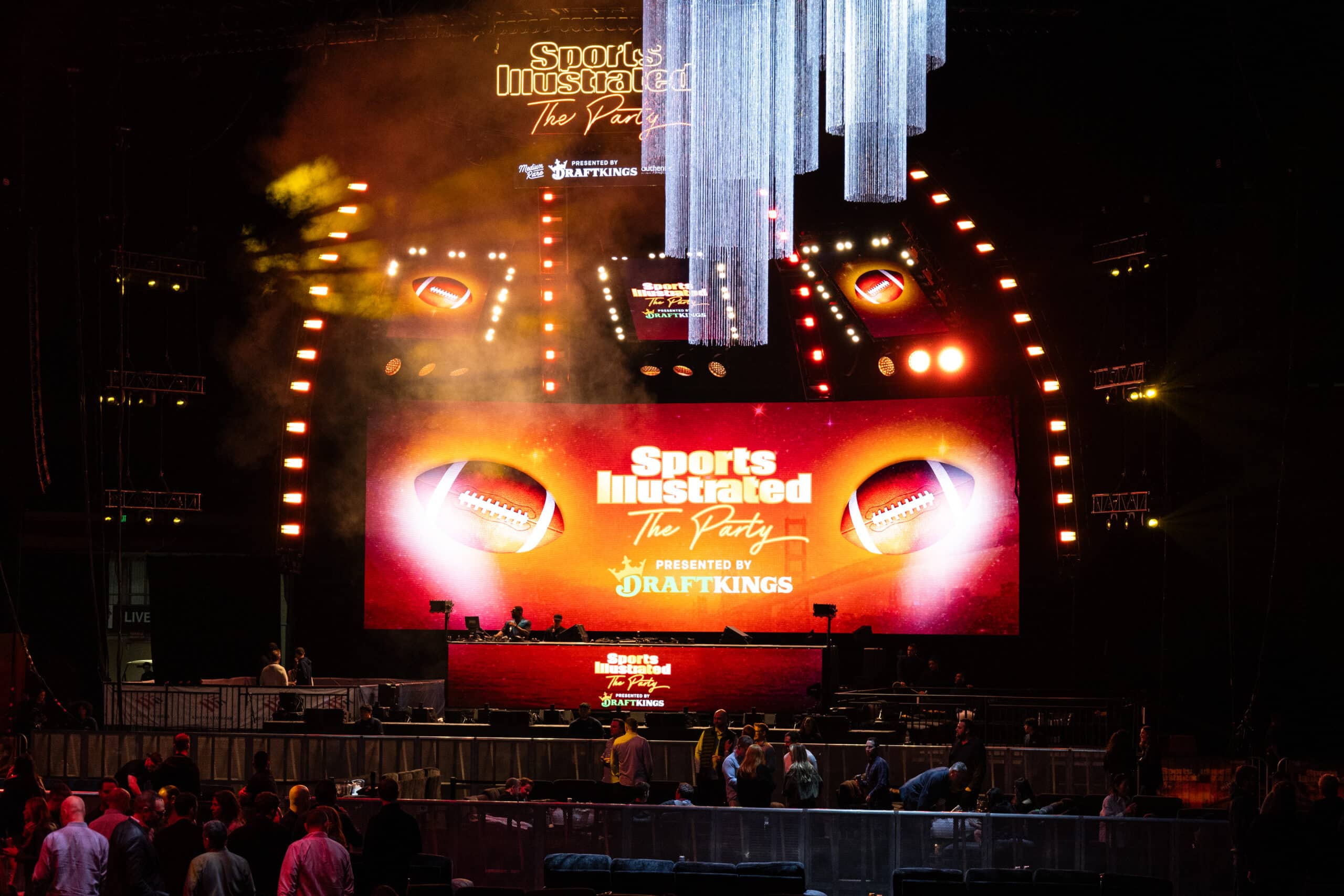 Sports Illustrated Super Bowl Party stage with vibrant lighting, large screen displaying "Sports Illustrated The Party" and football graphics, featuring a DJ booth and event attendees.