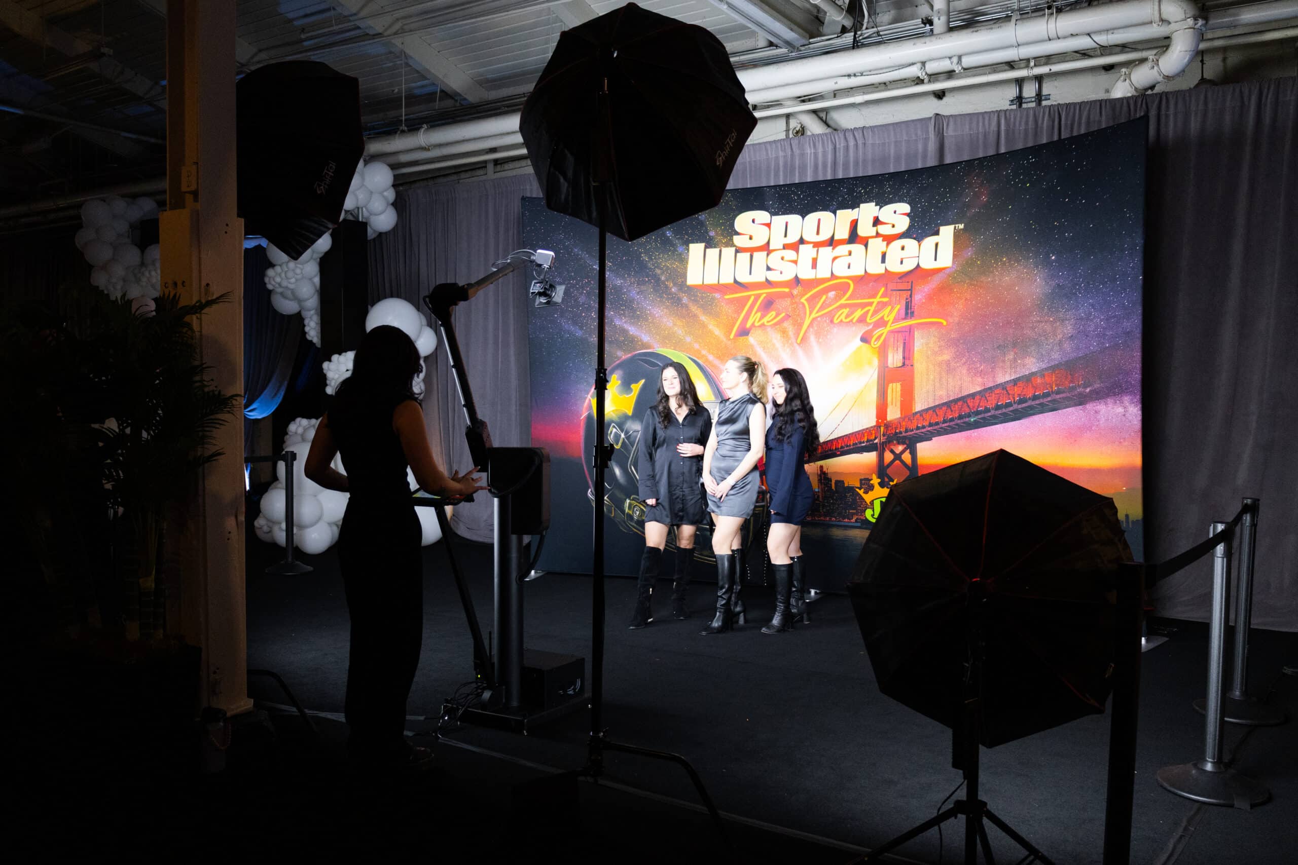 Three women posing for a photo in front of a vibrant backdrop featuring "Sports Illustrated The Party," with a camera setup and lighting equipment visible, highlighting the event's glamorous atmosphere.