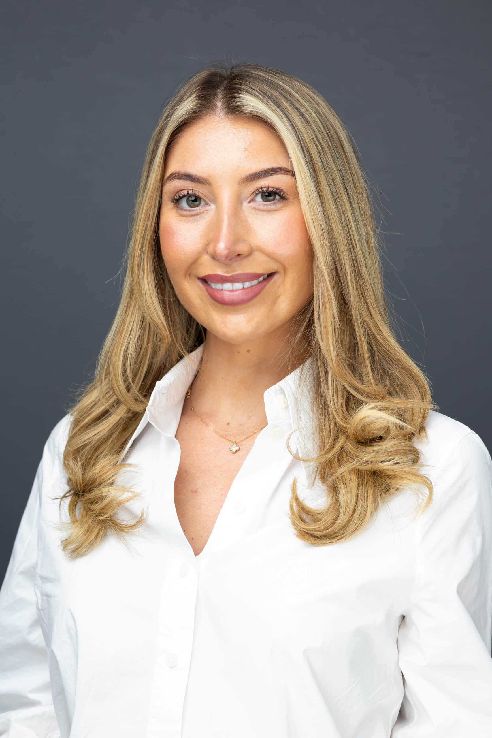 Professional woman with blonde hair wearing a white shirt and a delicate necklace, smiling against a gray background, representing high-quality headshot photography for corporate branding.
