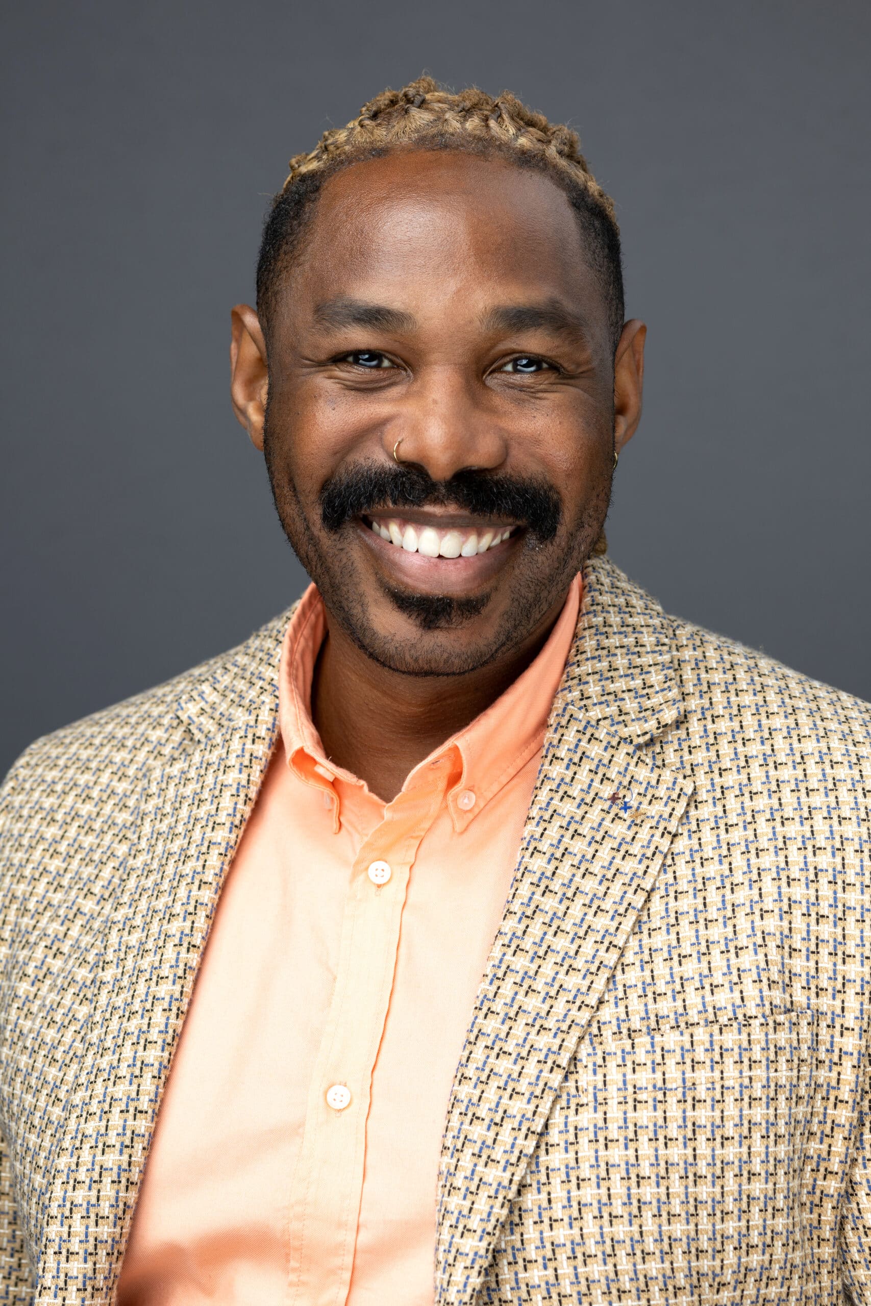 Professional male headshot featuring a smiling man with a mustache and nose ring, wearing a patterned blazer over an orange shirt, set against a neutral gray background, showcasing confidence and approachability for corporate branding.