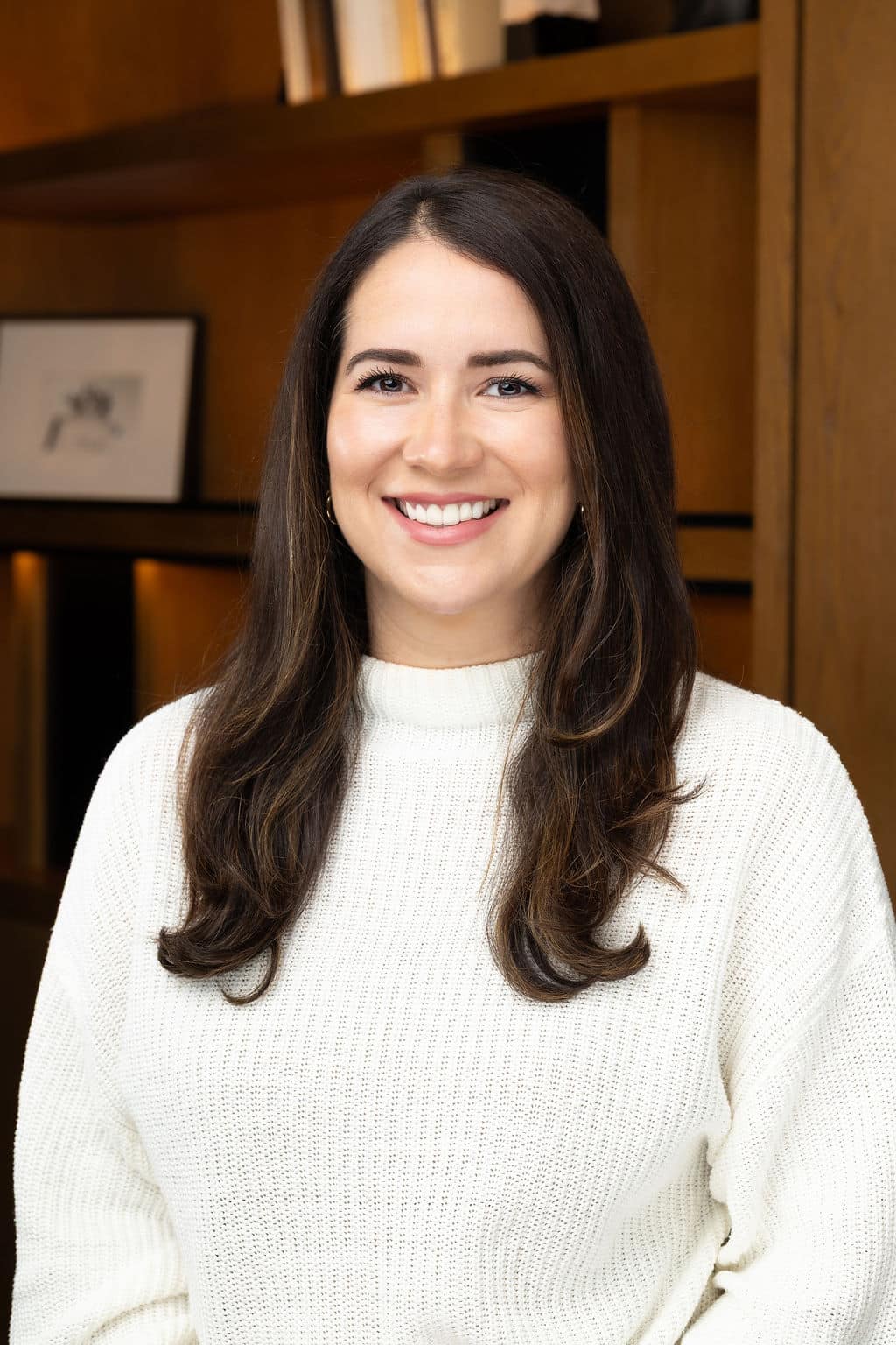 Professional woman smiling in a white sweater, showcasing a polished headshot against a warm, modern backdrop, reflecting confidence and approachability for corporate branding.