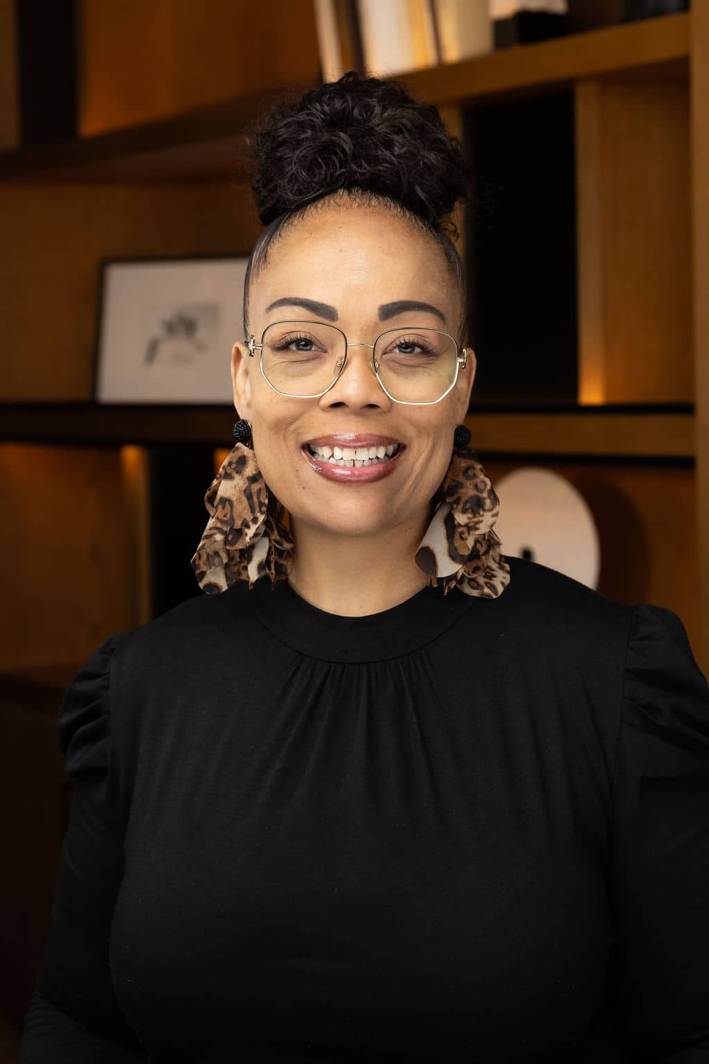 Professional woman smiling, wearing glasses and a black top, with stylish earrings, set against a warm-toned background, highlighting confidence and approachability in a headshot context.