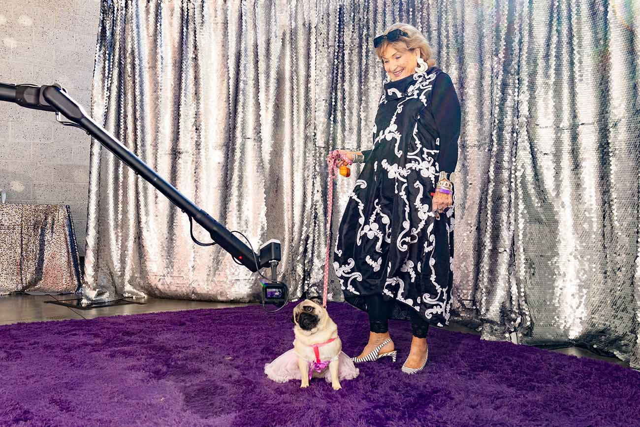 Woman in glamorous outfit with a stylish pug on a purple carpet, surrounded by a sequined backdrop and photo booth equipment at the Fur Ball 2025 event.