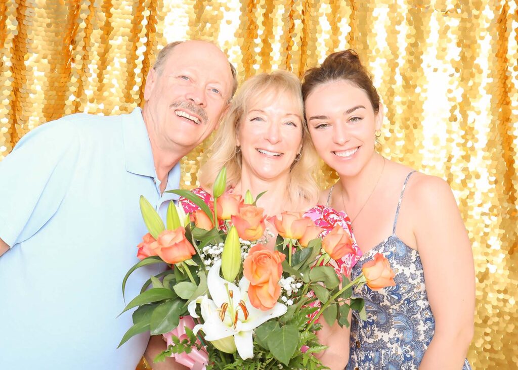 Three smiling individuals posing together in front of a shimmering gold sequin backdrop, holding a bouquet of orange roses and white lilies, representing a joyful moment at an event.