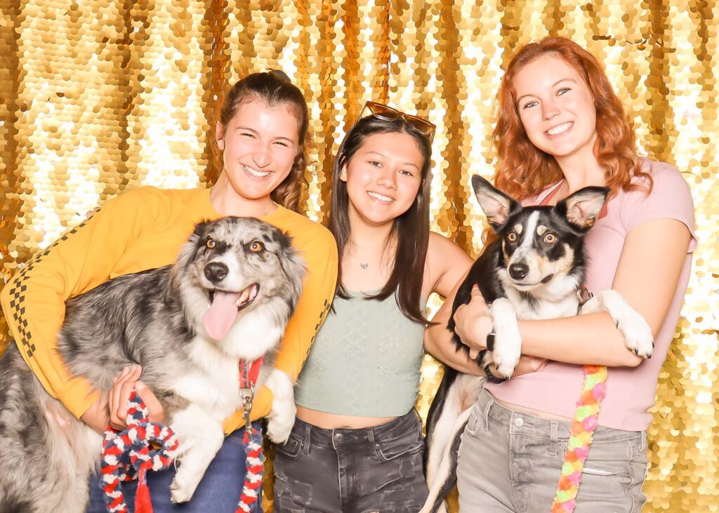 Three young women posing with dogs in front of a shimmering golden backdrop, showcasing a fun photo booth experience ideal for events.