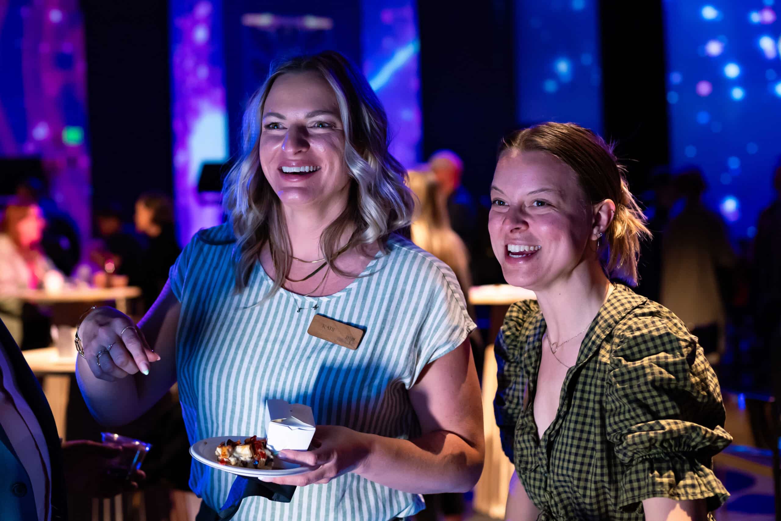 Two women enjoying food at a lively corporate event, smiling and engaging with guests, with colorful ambient lighting in the background, showcasing the vibrant atmosphere of the gathering.
