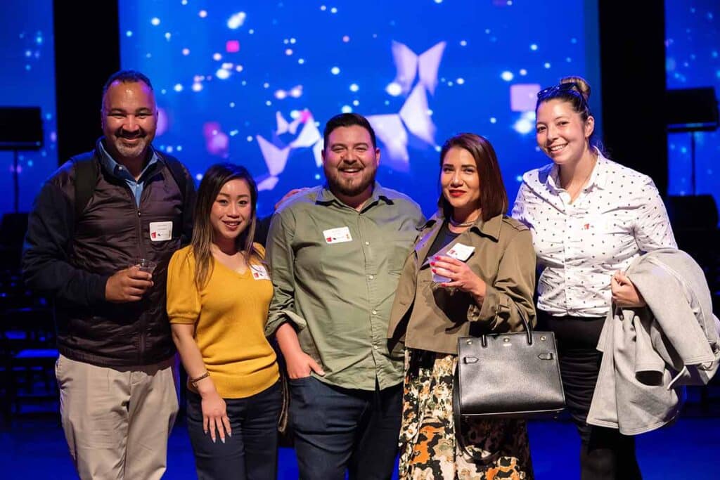 Group of five smiling individuals at a corporate event, showcasing event photography with vibrant background lighting, emphasizing brand engagement and social interaction.