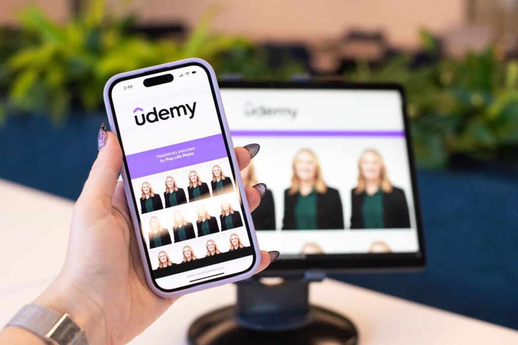 Hand holding smartphone displaying Udemy headshot gallery, featuring professional headshots by Pop Life Photo, with a monitor in background showing similar content, surrounded by greenery.