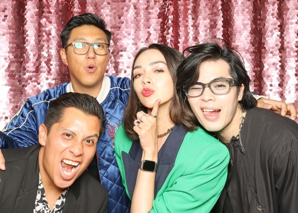 Group of four friends posing playfully in a photo booth with a shiny pink backdrop, showcasing fun expressions and stylish outfits, ideal for event photo booth inspiration.