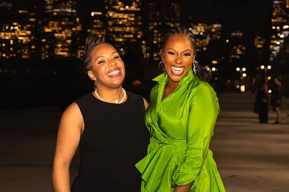 Two women smiling joyfully in elegant attire against a city skyline at night, showcasing the fun and engagement of photo booth experiences at corporate events.