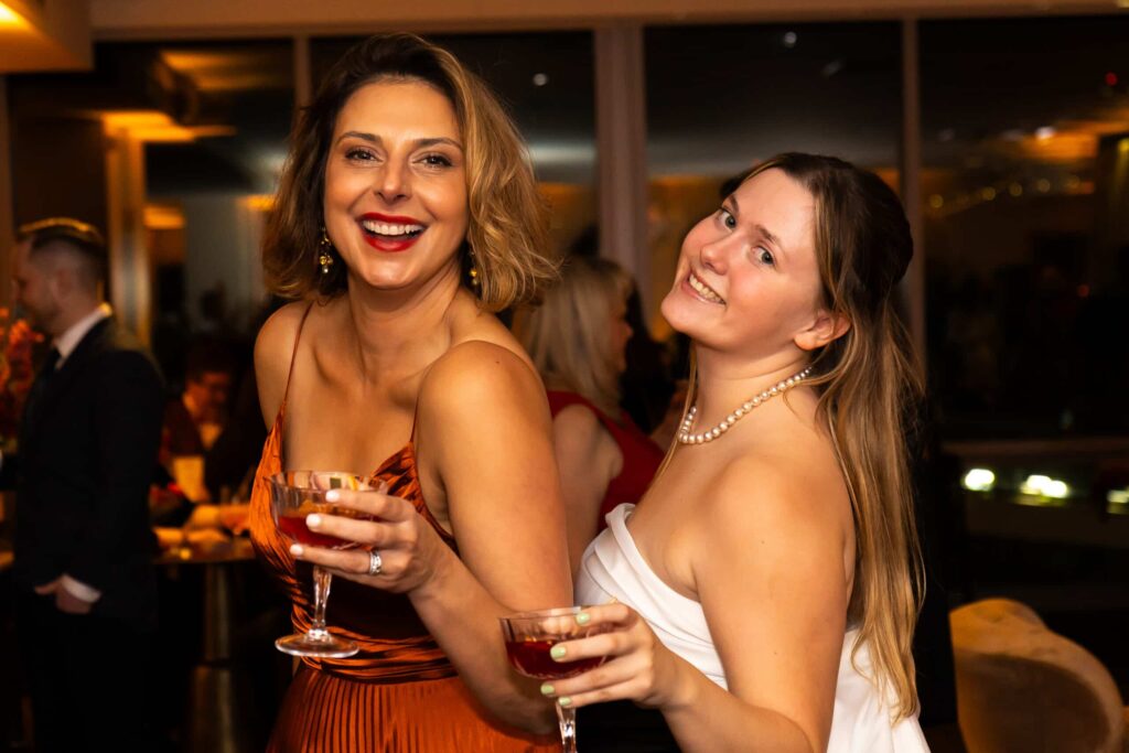 Two women at a holiday party, one in an orange dress and the other in a white dress, smiling and holding cocktails, with a festive atmosphere in the background.