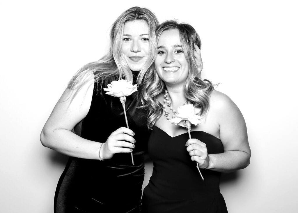 Two women posing together with white roses in a photo booth setting, celebrating an event, showcasing joyful expressions and stylish outfits.