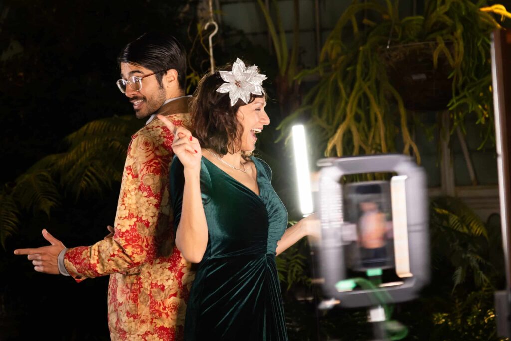 Couple posing back-to-back in stylish outfits at a graduation party photo booth, surrounded by greenery and lighting, capturing celebratory moments.