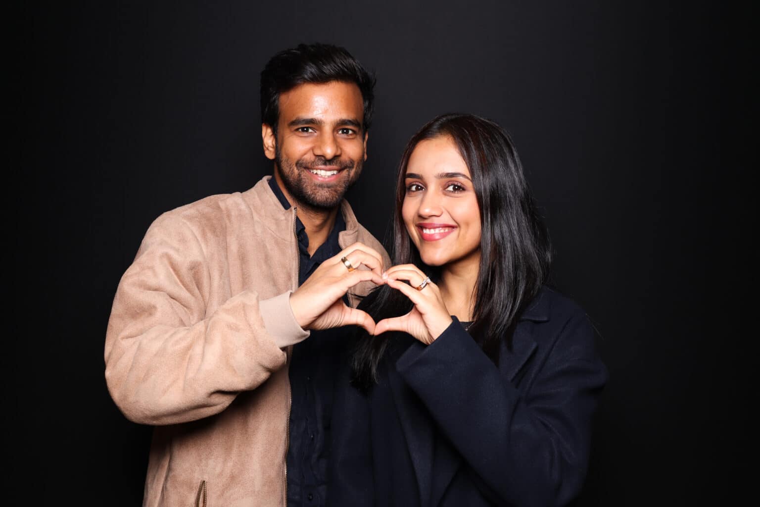 Couple smiling and making heart shapes with their hands against a black backdrop for a