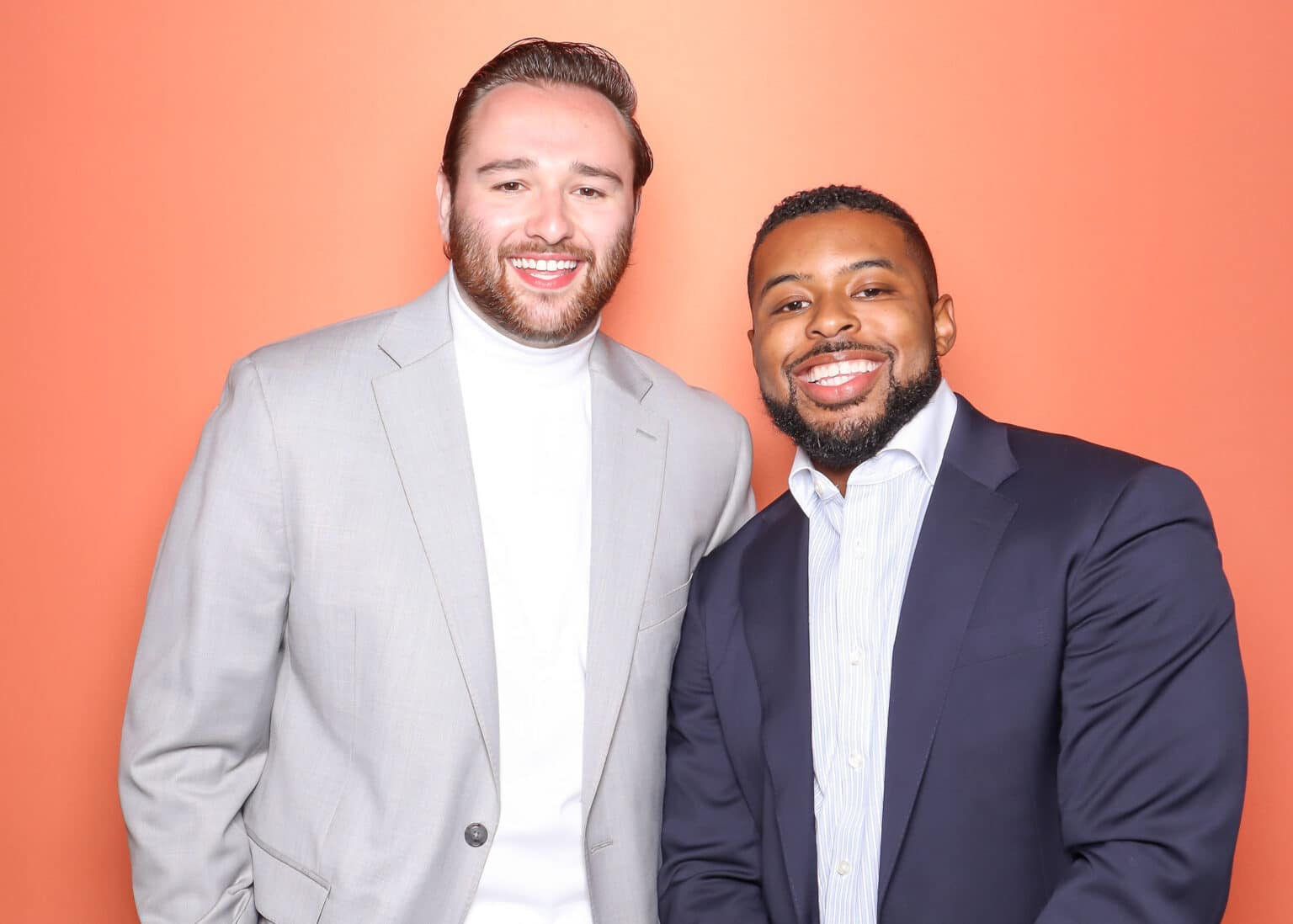 Fresh coral photo booth backdrop with two smiling men in suits.