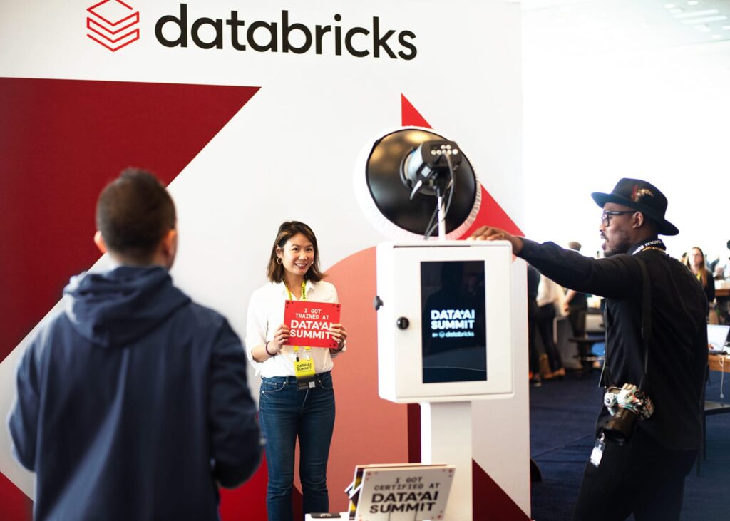 Woman holding a sign that says "I GOT TRAINED AT DATA+AI SUMMIT" in front of a Databricks photo booth, with attendees engaging in a corporate event setting.