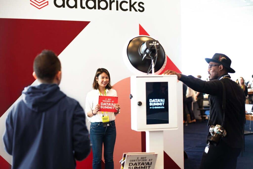 Photo booth at a tech event with a woman holding a sign that reads "I GOT TRAINED AT DATA AI SUMMIT," featuring a Databricks logo backdrop and attendees engaging with the booth.