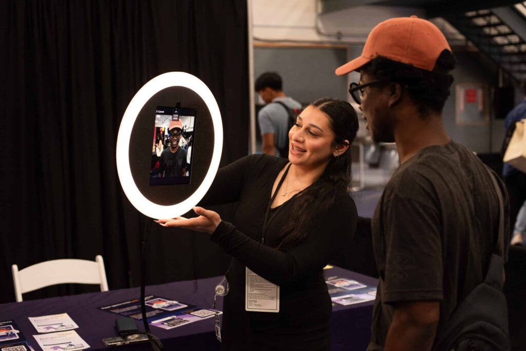 Woman demonstrating a roaming AI photo booth with a circular light, engaging with a man viewing his photo on the screen, event setting showcasing innovative entertainment technology.