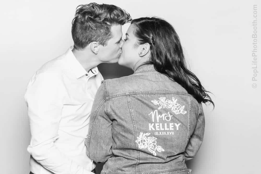 Couple kissing in photo booth, woman wearing "Mrs. Kelley" denim jacket, wedding celebration.