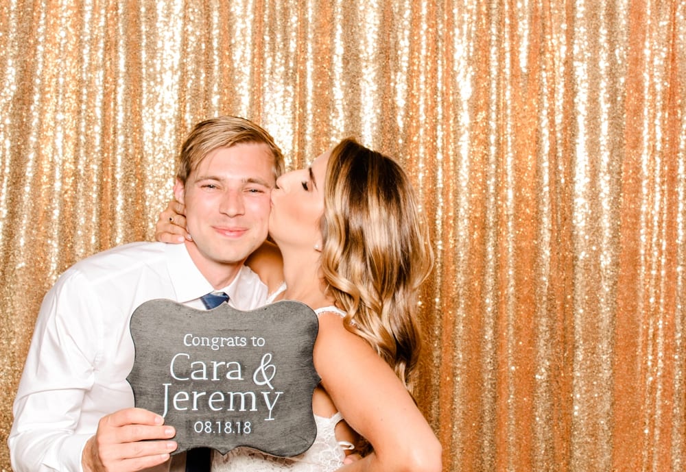 Couple celebrating with a sign reading "Congrats to Cara & Jeremy" in front of a shimmering gold backdrop at a wedding photo booth.