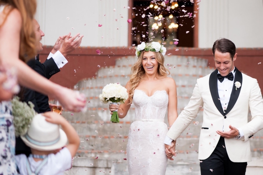 Bride and groom joyfully exiting wedding venue, surrounded by flower petals, with guests celebrating in the background, capturing a romantic spring wedding moment.