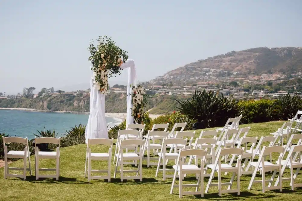 Outdoor wedding ceremony setup with white chairs and floral arch overlooking the beach and hills in California, ideal for event planning and venue selection.