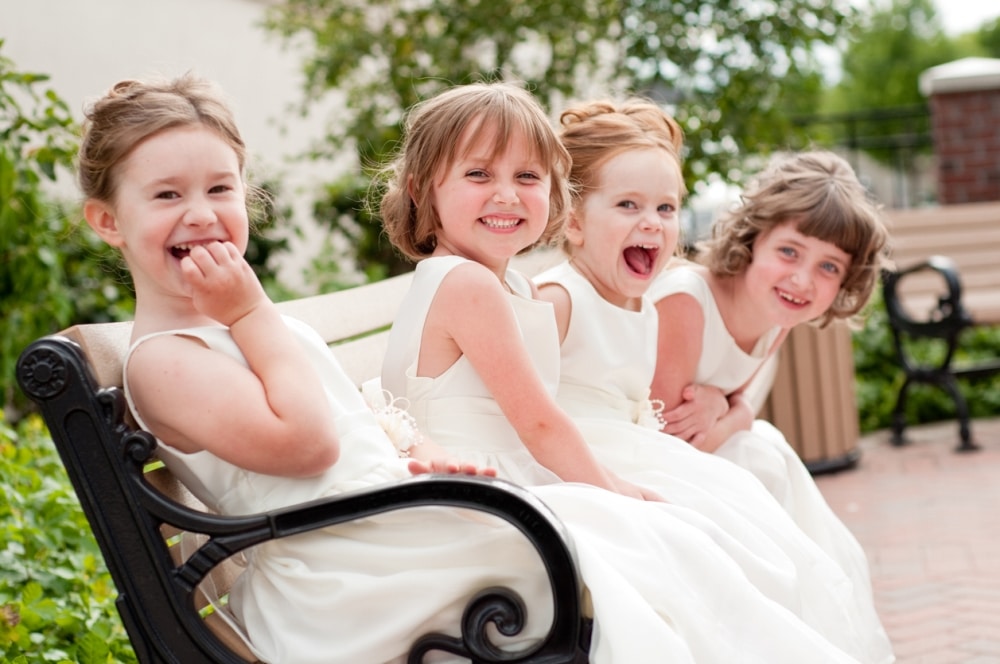Four young girls in white dresses joyfully sitting on a bench, smiling and laughing, capturing the essence of fun at a wedding celebration.