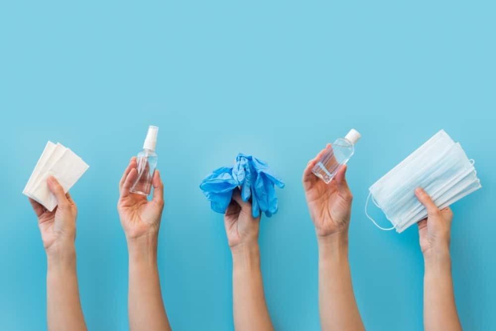 Hands holding sanitizing supplies: antiseptic spray, disinfectant wipes, blue gloves, and face masks, against a blue background, emphasizing safety measures for events during COVID-19.