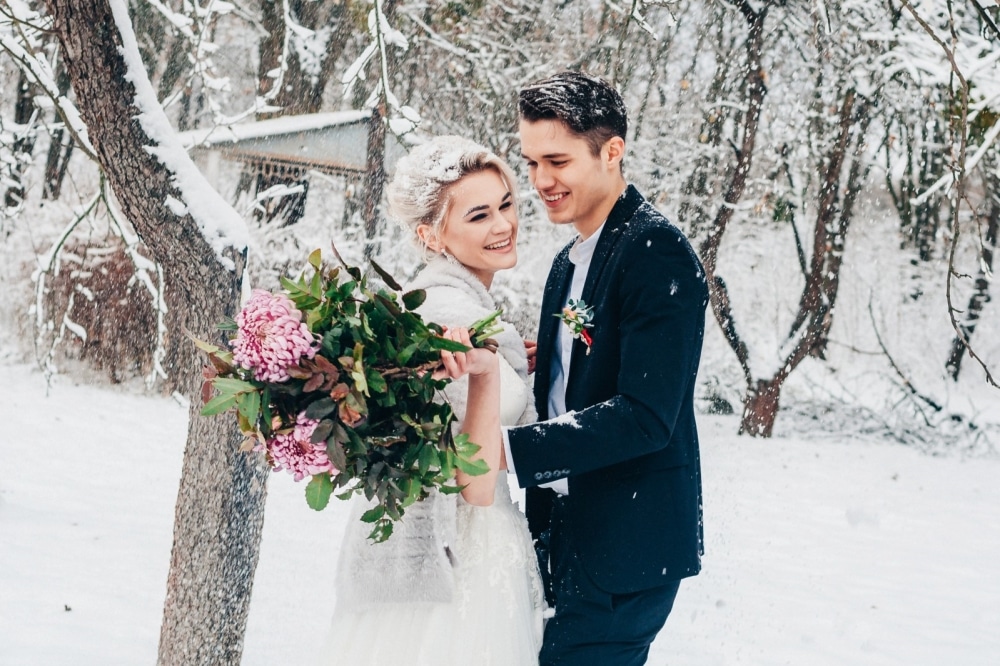 Bride and groom smiling in a snowy winter setting, holding a bouquet of flowers, embodying a romantic wedding moment.