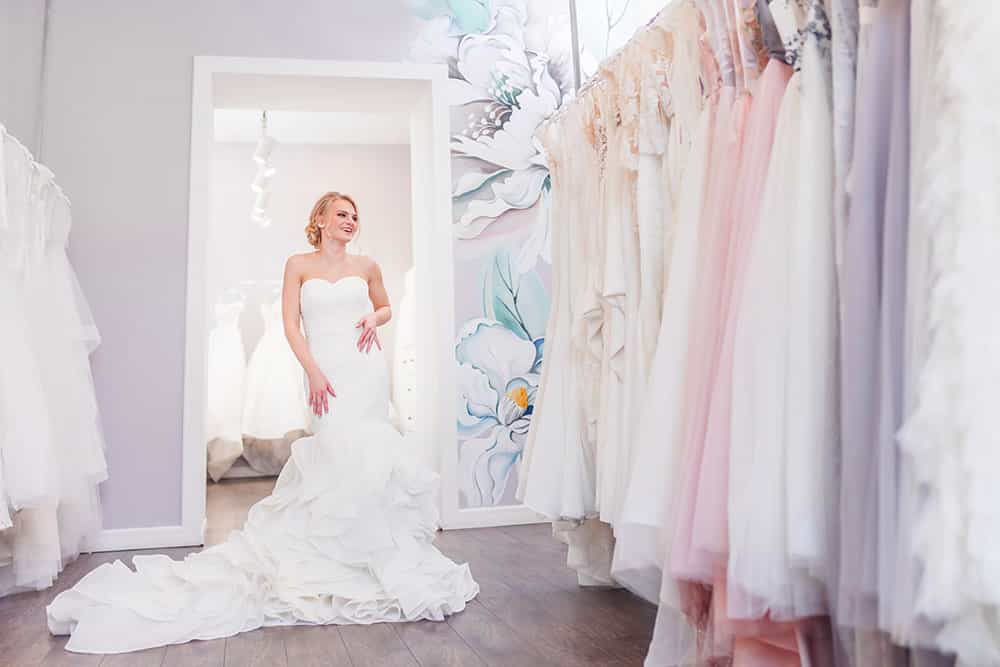 Attractive bride in elegant wedding dress smiling in bridal store surrounded by various wedding gowns.