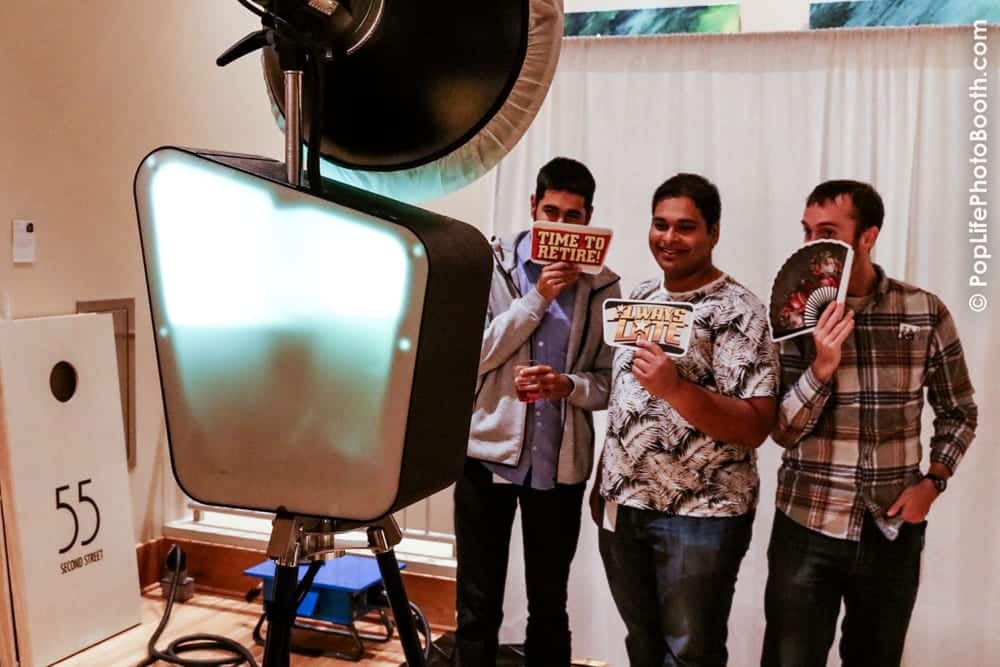 Friends enjoying a photo booth experience, holding playful props like "Time to Retire" and "Always Late," with a backdrop and lighting setup, relevant to event planning and photo booth rentals in San Francisco.