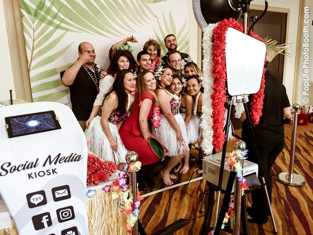 Group of people in festive attire posing at a photo booth with a "Social Media Kiosk," surrounded by tropical decorations, capturing a fun moment at a corporate holiday party.