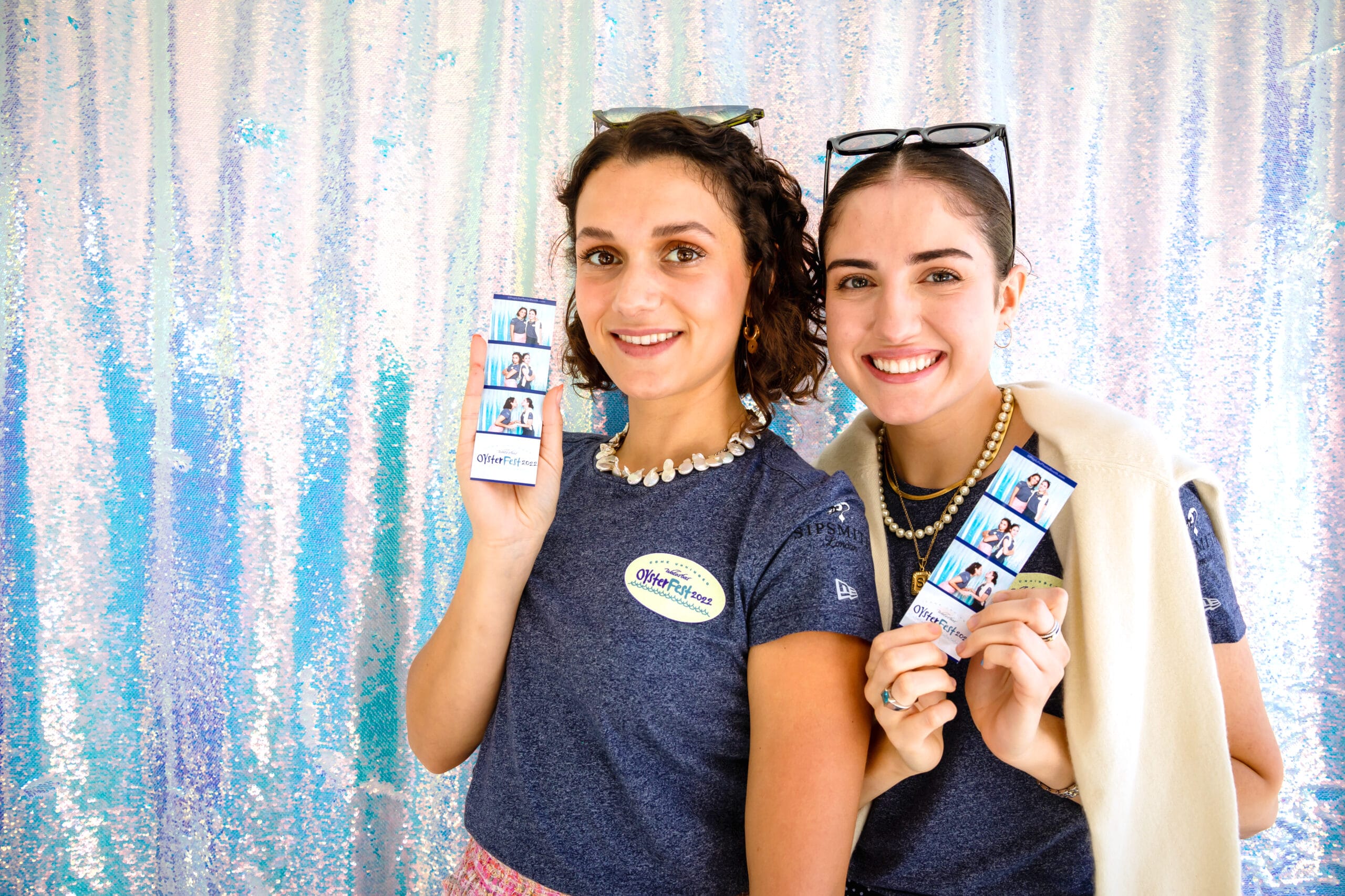 Two smiling women holding photo booth strips in front of a shimmering backdrop, promoting Pop Life Photo booth rental services for events in Sunnyvale.
