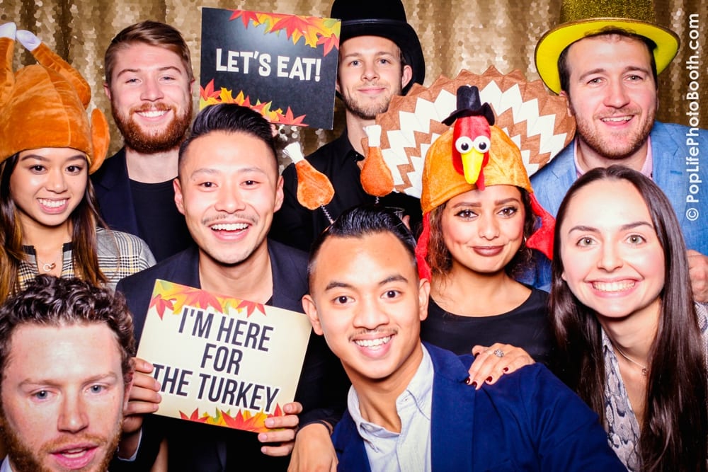 Group of friends celebrating Thanksgiving at Yelp party, wearing festive hats and holding signs "LET'S EAT!" and "I'M HERE FOR THE TURKEY," with a glittery backdrop.
