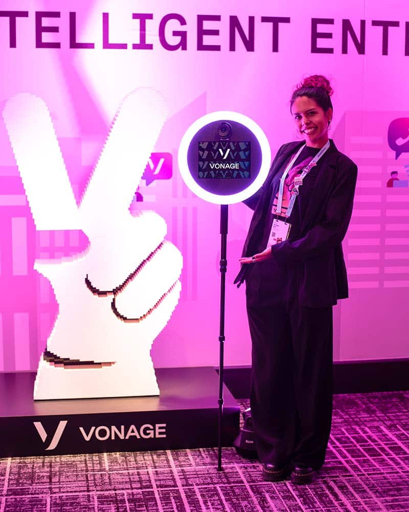 Woman posing with a photo booth setup featuring a ring light and a "Vonage" logo in a vibrant event setting, emphasizing photo booth marketing potential for brands.