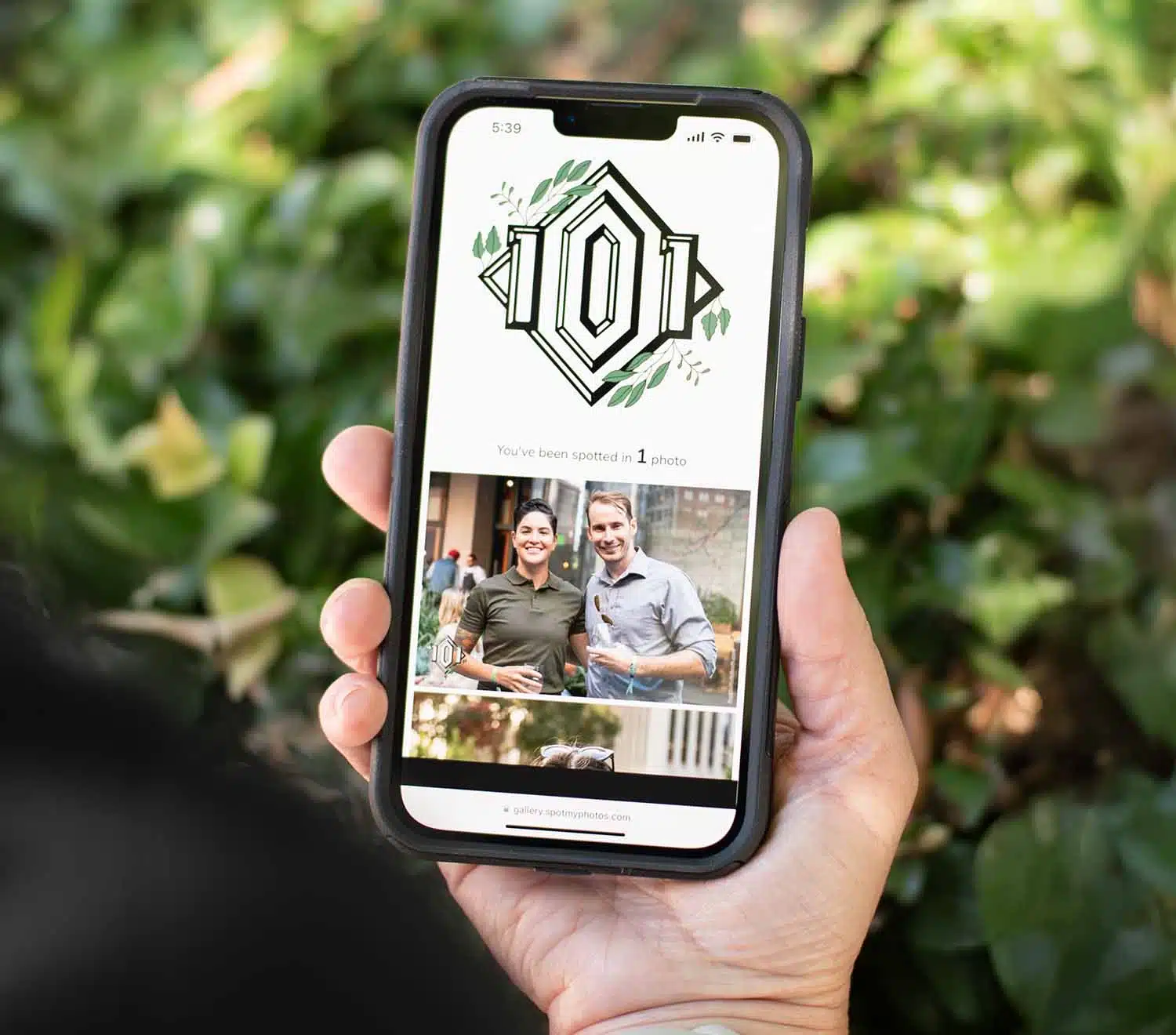 Person holding smartphone displaying a photo notification from an event, surrounded by greenery, emphasizing real-time engagement and sharing from Pop Life Photo's services.