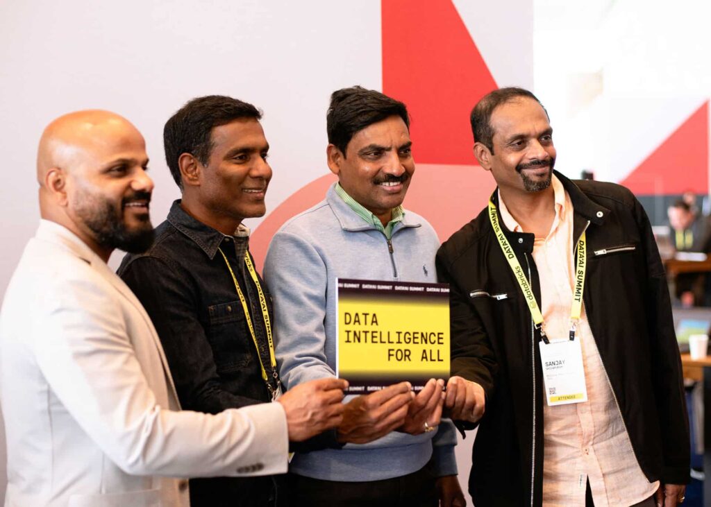 Group of four men posing together at a corporate event, holding a sign that reads "DATA INTELLIGENCE FOR ALL," showcasing camaraderie and engagement at a professional gathering.