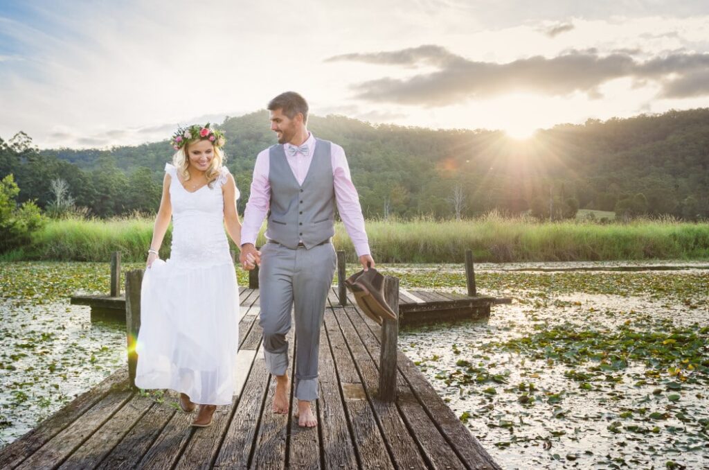 Couple walking hand in hand on a rustic wooden dock at sunset, dressed in wedding attire, surrounded by scenic nature, symbolizing romance and wedding planning.