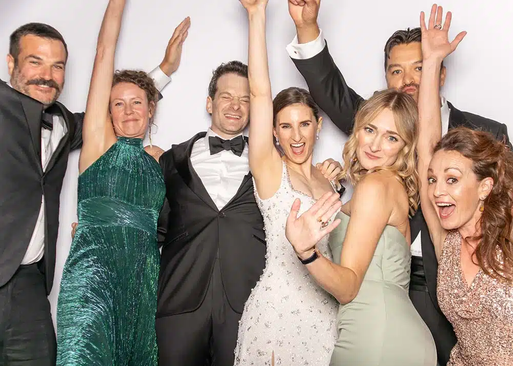 Group of seven smiling individuals in formal attire celebrating at a photo booth, showcasing joy and excitement, relevant to event marketing and entertainment trends for 2025.