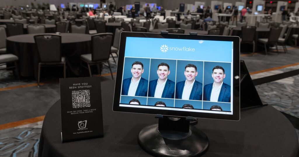 Photo booth display featuring a tablet with multiple headshot photos, promotional sign encouraging guests to scan a QR code for photo access, set in a spacious event venue with empty tables.