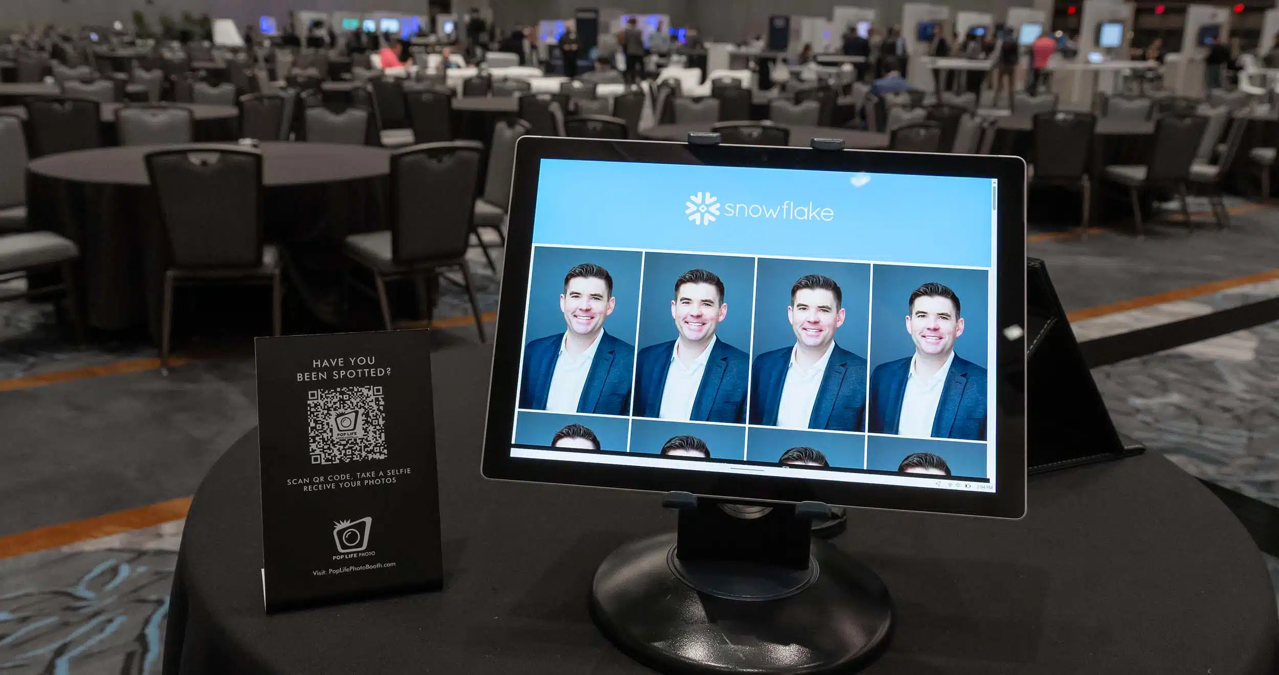 Photo booth display featuring multiple headshots on screen, with a QR code sign promoting Pop Life Photo booth services, set in a spacious event venue with tables and guests in the background.
