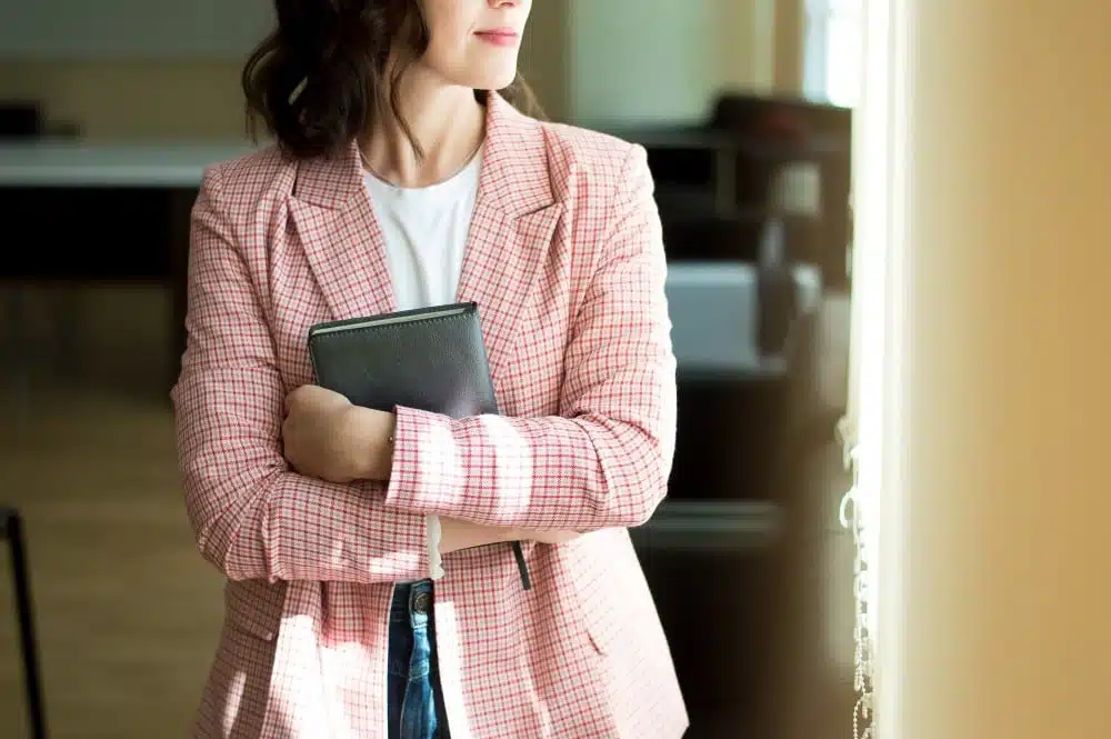 Woman in a pink checkered blazer holding a notebook, standing by a window in a modern event space, reflecting the theme of holiday event planning and organization.