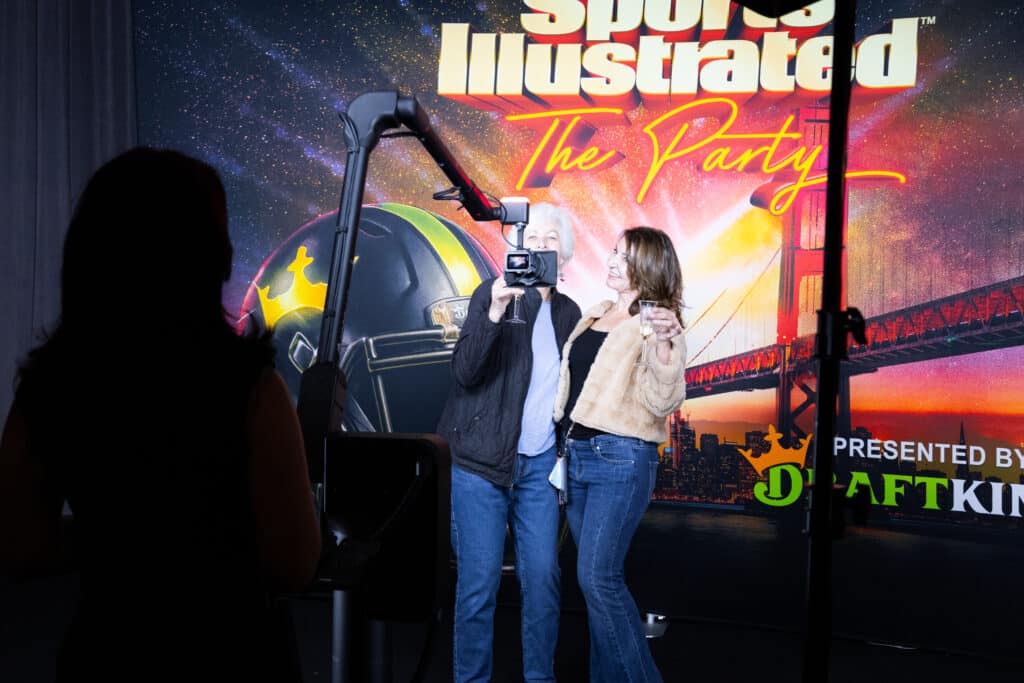 Two women posing for a photo at the Sports Illustrated Super Bowl Party, with a Glambot camera capturing the moment against a vibrant background featuring event branding and a football helmet.