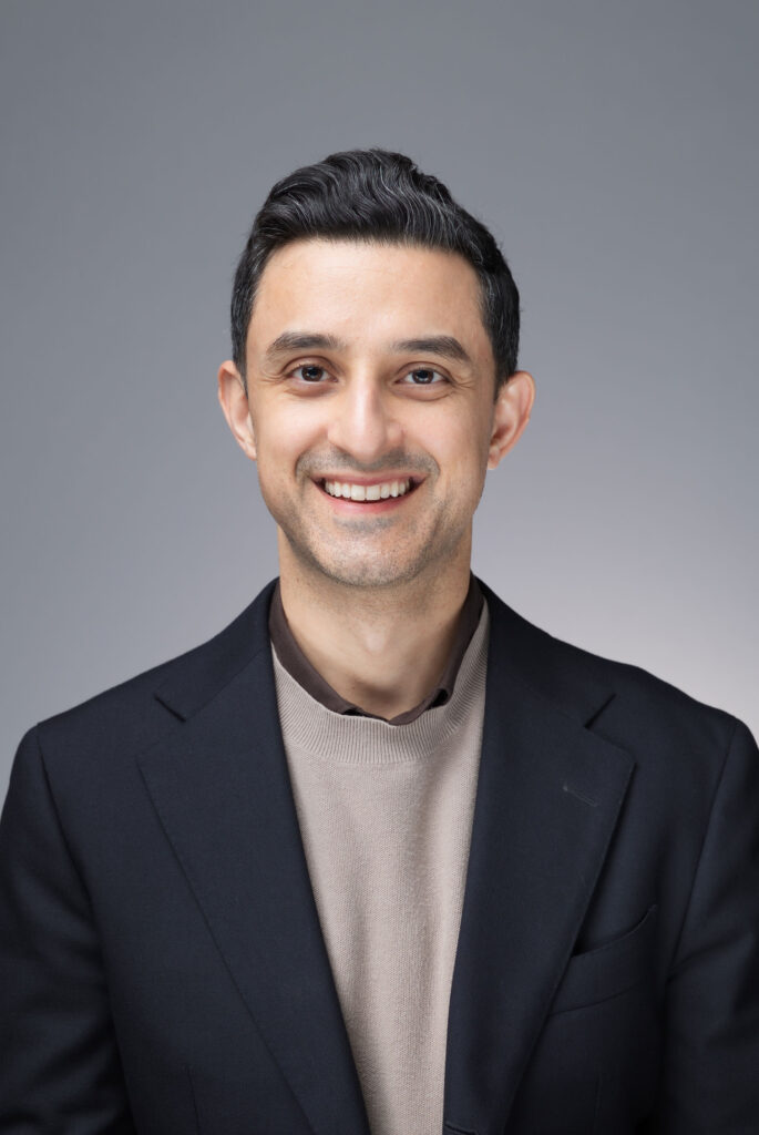 Professional headshot of a man in a dark blazer smiling against a neutral gray background.