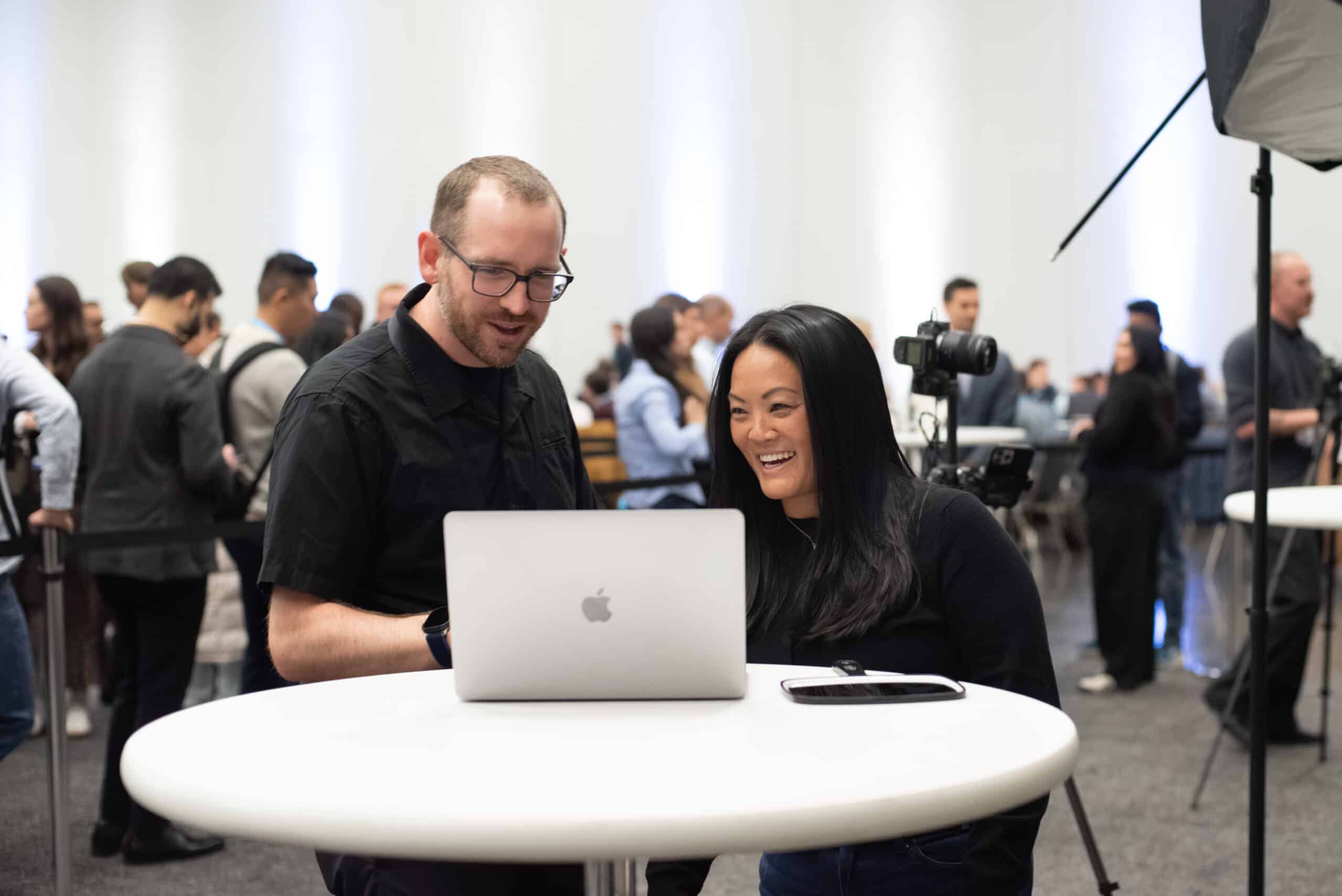 Two professionals engaging at a table, reviewing images on a laptop during the Snowflake AI Innovation Summit, with photography equipment and attendees in the background.